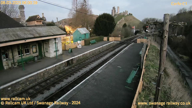 A view of a train station at dawn, featuring a stone building with a sloped roof and a green bench in front. To the left, there is a small green building. The platform is empty, with railway tracks running alongside it. In the background, a hill rises with remnants of ancient ruins atop it, silhouetted against the lightening sky. There are sparse trees nearby, and a wooden fence outlines the area, with some benches visible on the platform.