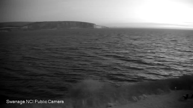 Black and white image of a calm sea with gentle waves. In the background, a low coastline can be seen, subtly outlining the land against the horizon. The sky appears cloudy and dim, suggesting either dawn or dusk. The atmosphere is tranquil, conveying a sense of peace.