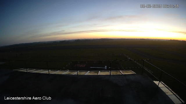 A panoramic view from a webcam at Leicestershire Aero Club, showing a grassy airfield under a pale blue sky at dawn. The horizon is lit with soft orange and yellow hues as the sun rises. In the foreground, there is a railing and part of a structure, while the airfield features a few white markers in the grass. The date and time in the corner indicate it is early morning.