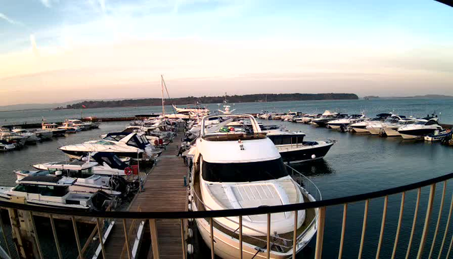 A waterfront scene showing numerous boats parked at a marina. In the foreground, several white and blue yachts are docked alongside a wooden walkway. The water is calm, reflecting the colors of the sky, which transitions from soft blue to warm orange as the sun sets. In the background, a distant green shoreline is visible. The overall atmosphere is peaceful and nautical.