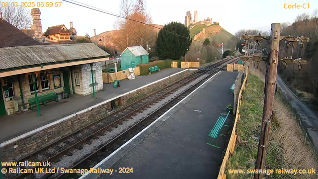 A quiet railway station with two sets of railway tracks running through it. On the left, there is a stone building with a sloping roof, featuring a green bench in front and a small green shed nearby. Several wooden benches line the platform, and a wooden fence separates the platform from a grassy area. In the background, a hill rises, topped with ruins of a castle. The sky is clear with warm morning light illuminating the scene.