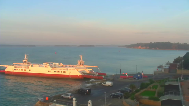 A ferry is docked at a harbor, with its ramp extended toward the shoreline. The water is calm, and there are a few boats in the distance. The scene is bathed in soft morning light, with a clear sky above. In the background, a shoreline with trees and hills can be seen, and there are several vehicles parked near the harbor, along with some green landscaping in the foreground.