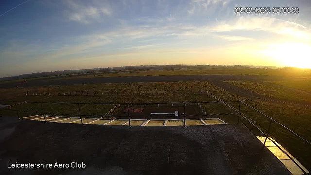 A panoramic view from a high vantage point over a grassy airfield at sunrise. The sky is mostly clear with soft clouds, and the sun is rising on the horizon, casting a warm glow over the landscape. In the foreground, there is a railing along a platform, and to the right, a paved runway stretches into the distance. Fenced areas are visible in the grass, suggesting designated zones on the airfield. The scene conveys a tranquil morning atmosphere at the Leicestershire Aero Club.