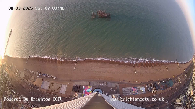 Aerial view of a beach at sunrise, with calm waves gently lapping the shore. In the foreground, there is a wide stretch of sandy beach that includes a few beach huts and attractions. A pier extends out into the sea from the left side of the image, while an old, partially submerged pier can be seen in the water further out. The overall scene is illuminated by soft morning light, creating a serene atmosphere. The bottom of the image displays some text indicating it is powered by Brighton CCTV, along with the date and time.