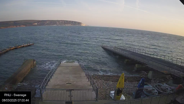 A view of the sea at sunrise, with gentle waves lapping against the shore. In the foreground, there is a concrete ramp leading down to the water, flanked by safety railings. To the right, several kayaks in blue and yellow are stored on a rocky beach. In the background, a wooden pier extends into the water, while cliffs are visible in the distance, partially illuminated by the early morning light. The sky is clear with a few wispy clouds, creating a serene atmosphere.