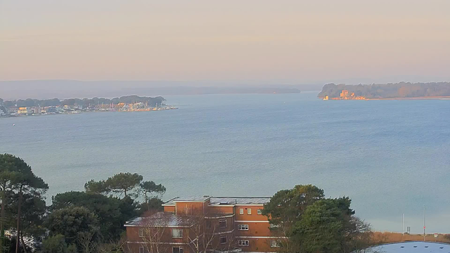 A panoramic view of a calm water body, with gentle waves reflecting soft colors of early morning light. In the foreground, there are a few clusters of green trees and a low brown building with multiple windows. To the left, a marina with various boats is visible along a shoreline, transitioning into a distant landscape featuring more greenery and a small, light-colored structure or island at the right side of the image. The overall scene feels serene and peaceful, under a clear sky.