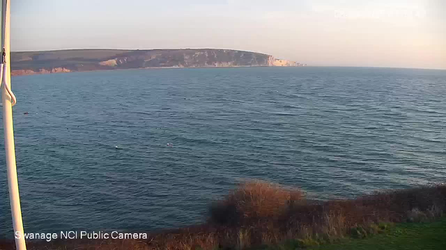 A coastal view shows calm blue water with gentle waves. In the distance, a rocky cliff juts out into the sea, partially illuminated by soft sunlight. The foreground features some brown grass and bushes along the water's edge, and the sky is a pale blue, indicating early evening.