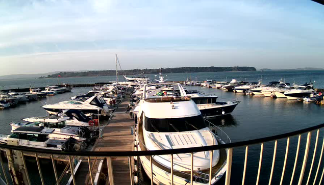 A wide view of a marina with numerous boats docked in the water. The foreground features a wooden walkway lined with several boats, including a large white motorboat at the center. In the background, the water extends towards a distant landmass with trees. The sky is partly cloudy, and the overall scene conveys a peaceful, recreational atmosphere.