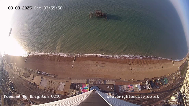 Aerial view of a sandy beach and the sea, captured from a high vantage point. The shoreline is visible, with gentle waves lapping against the sand. A partially submerged wooden pier extends out into the water. In the distance, the sunlight reflects off the ocean's surface. The foreground shows an amusement area with colorful patterns, while various structures are situated along the beach. The date and time are displayed at the top of the image, along with a logo for Brighton CCTV at the bottom.