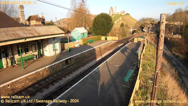 A train station view on a bright morning. The foreground shows a platform with several green benches and a signpost indicating "WAY OUT." To the left, there is a stone building with a sloping roof and a chimney. In the background, a hill is visible with a castle-like structure at the top. The scene is bathed in natural sunlight, casting long shadows on the ground. Train tracks run alongside the platform, leading off into the distance.