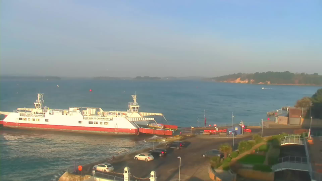 A calm waterfront scene featuring a large white and red ferry docked at a pier. The ferry is positioned near a road that has several parked cars. In the background, the tranquil blue waters extend towards distant land covered in trees, with a sandy cliff visible on the right side. The sky is mostly clear with soft lighting, indicating either early morning or late afternoon. Some buoys and a small boat are also present in the water.