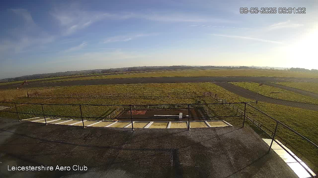 A wide view of a grassy field and a runway at Leicestershire Aero Club. In the foreground, there are several fences and a small area with red soil. The sky is clear with a few wispy clouds, and the sun is shining brightly, casting shadows on the ground. The scene is tranquil, suggesting a sunny morning.