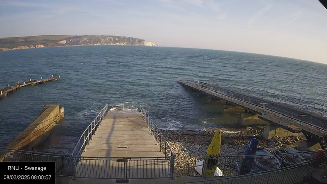 A view of a coastal area with a wooden pier extending into the water on the left and a stone quay leading down to the water in the center. Waves gently lap against the shore. To the right, there is another pier partially extending into the water. In the foreground, there are some kayaks positioned near the edge of the quay on the right, surrounded by pebbles. The background features a distant view of white cliffs under a clear sky. The scene conveys a calm, serene seaside atmosphere.