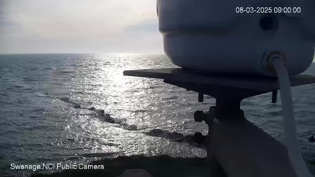 A view of the sea from a webcam, capturing a sunlit surface reflecting on the water. In the foreground, there is a portion of a white cylindrical object mounted on a platform. The horizon is visible in the distance, blending with a cloudy sky. The shoreline can be seen with rocks extending into the water. The overall scene conveys a calm, coastal atmosphere.