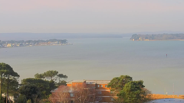 A scenic view of a calm body of water, with a mix of greenish and bluish hues. In the foreground, there are some trees and a brown building with multiple windows. The water is dotted with a few small boats, and a distant marina with several boats is visible on the left side. On the right side, a small island is partially visible with a structure on it. The background features a soft horizon with layers of trees and hills under a pale sky.