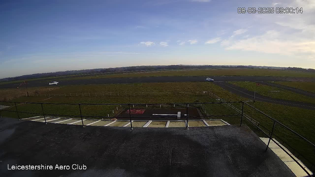 A wide open space at the Leicestershire Aero Club featuring a runway. In the foreground, there's a flat rooftop with a railing. Below, the runway stretches into the distance, lined by grass on both sides. A small white vehicle is seen driving along the runway. The sky is clear with a few clouds, and the overall scene is bright and sunny.