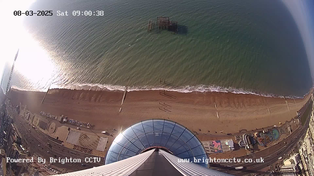 A high-angle view of a beach and ocean taken from a webcam. The image shows a sandy shoreline with gentle waves lapping at the sand, and a pier structure extends into the water. There are tracks on the beach, and a seaside promenade featuring amusement rides and various structures is visible nearby. The sky has a bright reflection on the water, indicating sunlight. The top of a glass dome is prominent in the foreground, suggesting the view is taken from a tall structure.