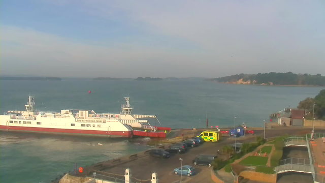 A view of a calm harbor with a large white and red ferry docked near a shore. The ferry has text indicating it is a chain ferry. In the foreground, there are several parked cars, including a bright green emergency vehicle. The water is a bluish color with distant islands visible on the horizon. On the right side, there is a landscaped area with grass and trees. The sky appears clear with a few clouds.