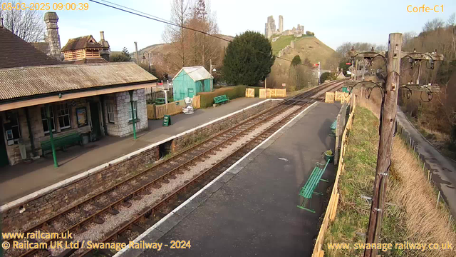 A view of a train station platform featuring an empty platform with wooden benches painted green. To the left, there is a stone building with a sloped roof and a chimney. In the background, there is a hill with ruins of a castle. Several trees surround the area, and a power pole with multiple wires stands on the right side of the image. The tracks run alongside the platform, and a fence can be seen in the distance near the end of the platform. The sky is clear and bright.