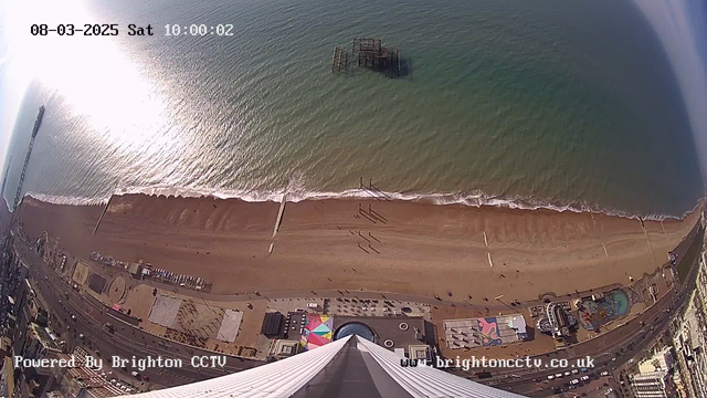 Aerial view of a beach and coastline. The scene includes a sandy beach meeting the ocean, with gentle waves rolling in. In the distance, an old, rusted pier structure is partially submerged in the water. A promenade runs along the beach with a few people walking. Colorful beach chalets and attractions can be seen near the shore, while vehicles are parked along the road adjacent to the beach. The image also shows the shadows cast by objects and structures, along with text displaying the date, time, and a logo at the bottom.