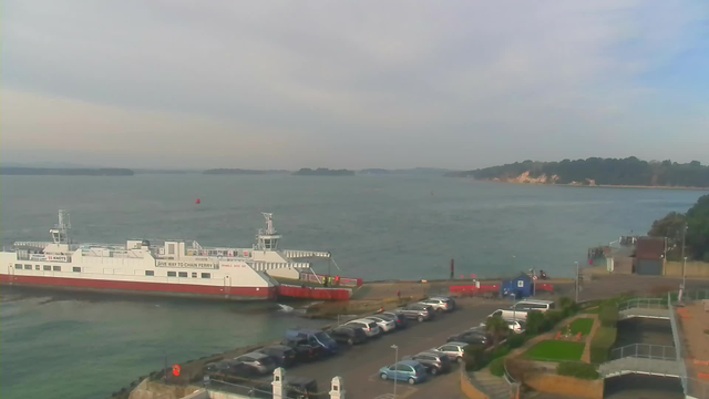 A ferry is docked at a terminal near a body of water, with several parked cars visible in the foreground. The water appears calm, and there are some small islands in the distance. The sky is mostly cloudy, with a hint of blue visible. On the right, there are some buildings and greenery, while a red buoy is floating in the water nearby.