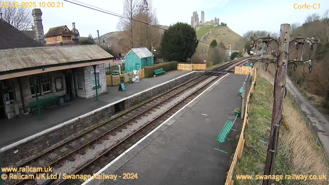 A view of a railway station taken from a webcam. The foreground features a platform with green benches and a stone wall. To the left, there is a building with a sloped roof and several windows. Behind it, a small green shed is visible. In the background, a hill rises with ruins of a castle at its summit. Railway tracks are visible running through the scene, and a pole with electrical wires stands near the edge of the platform. The sky is predominantly clear with some clouds.