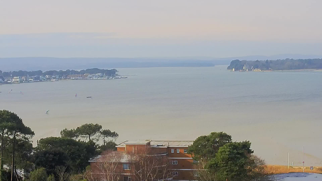 A panoramic view of a calm body of water stretches across the image, reflecting subtle shades of gray and blue. In the foreground, there is a low-rise building made of orange-brown brick, partially obscured by green trees. To the left, a marina with several boats is visible, along with distant sailboats on the water. On the right side, an island can be seen, featuring lush greenery and a notable structure, possibly a small lighthouse or building. The sky is light with soft clouds, creating a serene atmosphere.