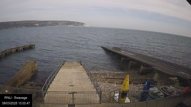 A view from a webcam showing a calm sea with gentle waves and a cloudy sky. In the foreground, there is a concrete ramp leading down to the water, flanked by a railing. To the right, two boats in bright yellow and blue sit on the shore next to small rocks. In the background, a long pier extends out into the sea, and a cliffside can be seen in the distance. The date and time are displayed at the bottom left corner of the image.