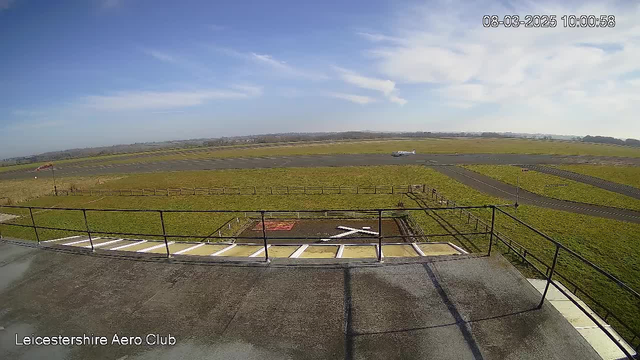 A clear blue sky with some scattered clouds over a grassy airfield. In the foreground, a low railing borders a flat rooftop area with white markings. In the background, a small airplane is taxiing on the runway. The ground is a mix of green grass and gray asphalt, with several grassy areas and wooden fences visible in the distance. The time displayed is 10:00 AM on March 8, 2025.