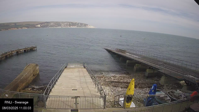 A coastal scene featuring calm waters with a distant shoreline. In the foreground, stairs lead down to a rocky area near the water's edge. To the right, there are two kayaks on display: one is yellow and the other is blue. A wooden pier extends into the sea, with a railing along its sides. The background shows a clear sky with some clouds and cliffs in the distance. The time and location information is displayed in the lower left corner.