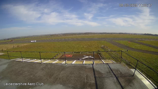 A wide view of a grassy airfield under a bright blue sky with a few scattered clouds. In the foreground, metal railings are visible with the edge of a flat rooftop. On the airfield, there are taxiways and a small white aircraft positioned nearby. A red and white windsock stands to the left. The background shows a horizon with distant hills. The scene appears to be well-lit and clear.