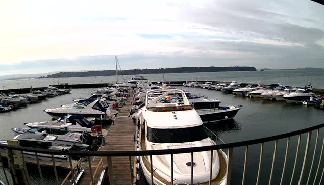 A scenic view of a marina filled with numerous boats docked along a wooden walkway. In the foreground, there is a large white boat with a dark windshield. The water is calm, reflecting the cloudy sky above. Several smaller boats are moored alongside, and the distant shoreline features trees and possibly hills. The image conveys a tranquil, marine atmosphere with various vessels arranged neatly.