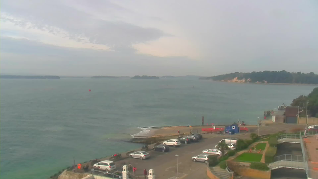 A coastal view featuring calm waters and several islands in the distance under a slightly cloudy sky. In the foreground, there is a parking lot with numerous cars, as well as a grassy area with some people present. A small harbor structure is visible to the right, alongside buildings and some construction equipment nearby. A red buoy floats in the water, indicating navigation points.