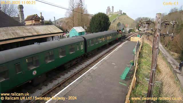 A green steam train is standing at a railway station platform with several carriages visible. The station features a stone building with a sloped roof and a tower, surrounded by trees. In the background, a hill rises with the ruins of an old castle. There are benches on the platform, and a person walks along a path beside the railway with a dog. The sky is clear, and the scene has a peaceful, rural atmosphere.