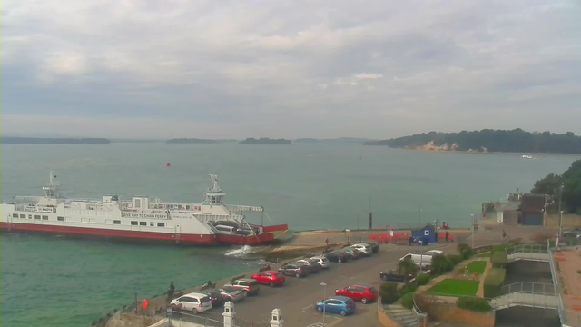 A large white and red ferry is docked at a port, surrounded by calm water. In the foreground, there is a parking area filled with several cars of different colors, including red and white. Beyond the ferry, a distant shoreline with trees and a sandy beach can be seen, under a cloudy sky. The scene captures a tranquil coastal setting.