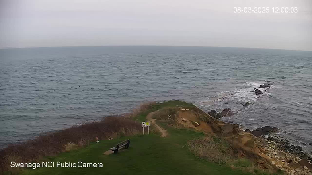 A coastal scene showing a grassy area leading to the edge of a cliff. There is a bench facing the sea, near a signpost with safety information. The water is calm, with gentle waves lapping against the rocky shoreline. The sky is overcast, creating a soft, diffused light over the landscape.