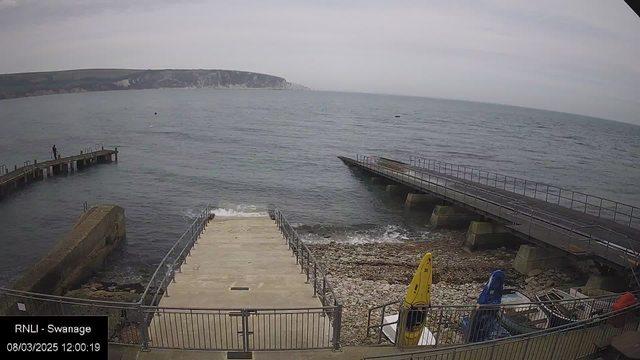 A coastal scene featuring a calm sea under a cloudy sky. There is a ramp leading down to the water, surrounded by a rocky shoreline. A wooden pier extends into the sea on the left, where a person is standing. On the right, a small boat ramp is visible with several boats nearby, including yellow and blue kayaks. The background shows cliffs and hills along the coastline.