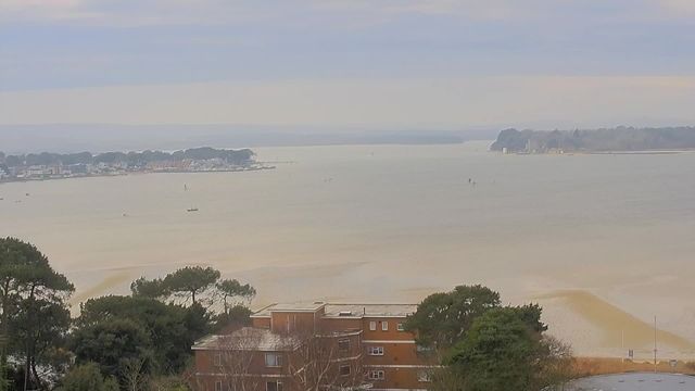 A panoramic view of a calm body of water, with soft waves lapping at the shore. The scene features a mix of brown sandy areas and darker patches of water. In the background, there is a distant shoreline lined with trees and buildings. Several small boats are visible on the water, some closer to the shore and others further away. The sky is light gray with hints of blue, suggesting a cloudy day. In the foreground, there are trees and a building with a reddish-brown exterior.