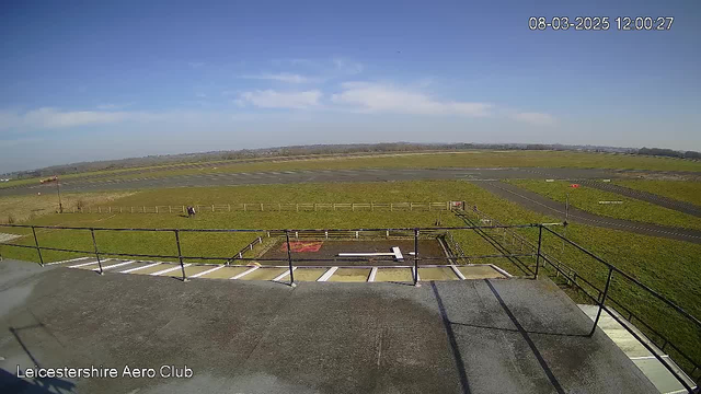 A wide view of an airfield under a clear blue sky. The grassy ground is visible with several sections of tarmac or runway intersecting. A fenced area can be seen in the center, with a white rectangular pattern on the ground. In the distance, low hills are faintly visible. A wind sock indicates the direction of the wind, and there are open spaces around the field, emphasizing the vastness of the area.