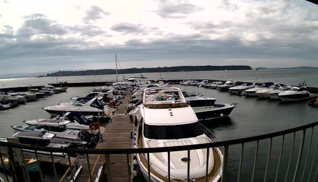 A view of a marina during the day, featuring numerous boats docked in the water. The foreground includes various motorboats and yachts, with some positioned on a wooden dock. The background shows a calm body of water under a mostly cloudy sky, with a distant shoreline visible. A small flag is seen on the pier, and several boats are aligned in the marina, creating a busy yet serene atmosphere.