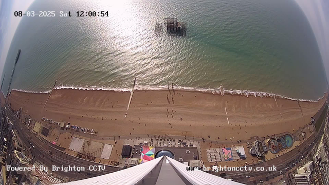 An aerial view of Brighton beach and seafront captured from a high angle. The image shows the sandy beach stretching below with a few people walking. Gentle waves lap at the shoreline. In the water, a derelict pier structure is visible. The beach area features colorful beach huts and various attractions, including a ride and a circular pool. The image has a timestamp indicating the date and time, and it is marked as powered by Brighton CCTV. The sky is mostly clear with some light clouds.