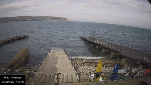 A coastal view showing the sea with small waves and a cloudy sky. In the foreground, there is a concrete ramp leading down to water, surrounded by metal railings. A rocky shoreline is visible next to the ramp. To the right, there are two brightly colored kayaks, one yellow and one blue, positioned near the edge of the ramp. In the background, cliffs can be seen in the distance along the coastline.