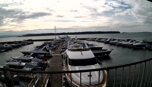 A view of a marina with numerous boats docked in the water. The foreground features a large white yacht near a wooden walkway, leading to smaller motorboats and sailing craft. The harbor is surrounded by calm water, reflecting a cloudy sky with patches of light. In the distance, there are hills and a shoreline, suggesting a serene and scenic environment.