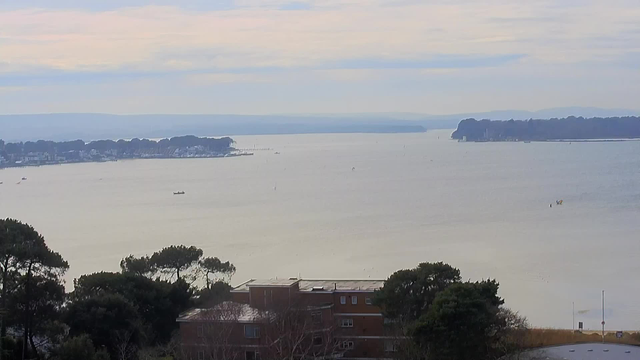 A panoramic view of a calm bay under a cloudy sky. The water appears smooth and is reflecting the muted colors of the sky. On the left side, there is a small town with buildings and a wooden pier extending into the water. Some boats are visible in the bay, and distant trees outline the shore on both sides. In the foreground, there are trees and rooftops visible, contributing to the natural landscape.