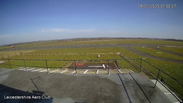 A view from a webcam at the Leicestershire Aero Club, showing a wide expanse of grassland and a tarmac runway. The sky is clear with few clouds, and the sun is shining brightly. In the foreground, there is a railing along a rooftop with a view of the runway. A small white structure is visible on the ground, possibly a landing area or signal. A solitary figure is seen walking on the grass near the edge of the runway. In the distance, there are trees and fields extending towards the horizon. The image is time-stamped, showing the date and time at the top right corner.