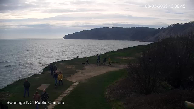 The image shows a coastal scene with a rocky shoreline and a calm sea under a cloudy sky. In the foreground, several people are walking along a grassy pathway, with some visible near a dirt path lined by stones. The landscape includes green grass and bare bushes. In the distance, cliffs rise along the coastline. The overall atmosphere is serene, indicative of an overcast day.