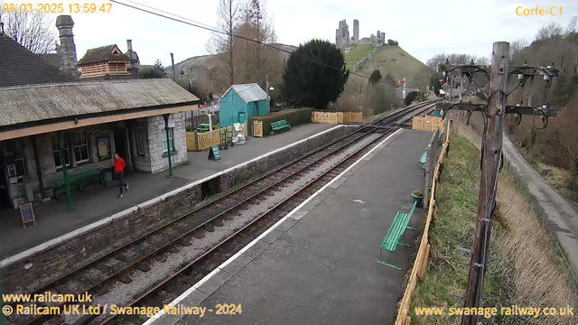 A railway station scene with a stone building featuring a roof and a canopy over the platform. In the foreground, a person in a bright red jacket jogs along the platform beside green benches. To the left, there is a teal-colored shed and a small sign that reads "OUT." In the background, a hill rises with a castle ruin atop it. The railway tracks run parallel to the platform, disappearing into the distance on the right side. There are power lines and wooden poles on the right edge of the image. The scene is set under a cloudy sky.