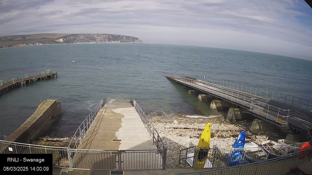 A view of a seaside area showing a wooden dock extending into blue water. In the foreground, there is a concrete ramp leading down to the water, flanked by a rocky shoreline. Two kayaks, one yellow and one blue, are placed on the ground beside the ramp. In the background, cliffs and hills rise along the shore under a partly cloudy sky. The scene conveys a calm, coastal atmosphere.