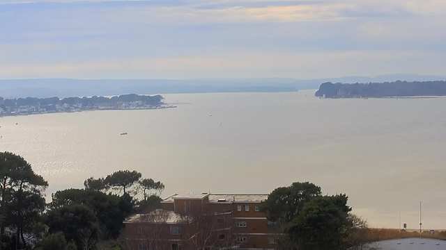 A panoramic view of a calm water body reflects soft light under a cloudy sky. In the distance, a shoreline with buildings and trees lines the water's edge. A small boat is visible on the water, with hints of distant hills beyond. The foreground features greenery and a low building, partially obscured by trees.