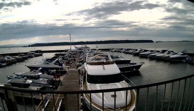 A view of a marina featuring numerous boats docked in water. In the foreground, a large white boat is prominent, with several smaller boats surrounding it. The scene is set under a cloudy sky, with rippling water and a distant shoreline visible in the background. The docks are made of wood and there are boats of various sizes, some with colorful accents, moored along the sides.